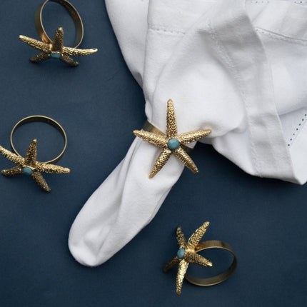 Casa Duniya's Gold and Turquoise Starfish Napkin Rings on a White Napkin and Blue Background