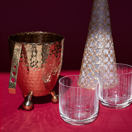 Casa Duniya's Gold Chameli Hammered Gold Ice Bucket with two whiskey glasses, a decorative gold christmas tree on a red background